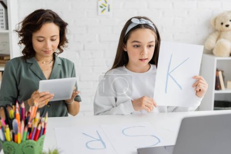 Preteen girl holding paper with letter k while logtherapy online lesson on laptop near mom at home 