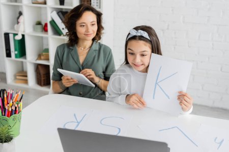 Lächelndes Mädchen mit Papier und Brief während Logopädie-Unterricht auf Laptop in der Nähe von Mama mit digitalem Tablet zu Hause 