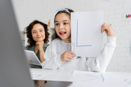 Child sticking out tongue and holding paper with letter during speech therapy lesson on laptop near mom at home 