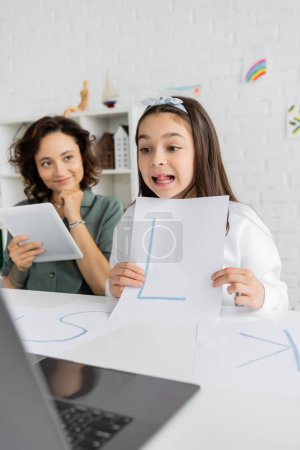 Niña sosteniendo papel con carta durante la lección de video de terapia del habla cerca de mamá sonriente con tableta digital en casa 