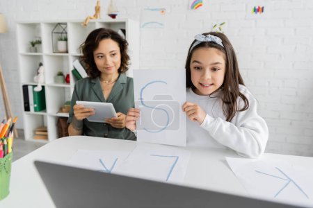 Niño preadolescente sonriendo y sosteniendo papel con carta durante la lección de terapia del habla en la computadora portátil cerca de los padres en casa 
