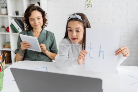 Preteen child holding paper with letters near laptop and mom during online logopädie at home 