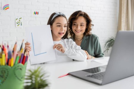 Smiling woman looking at cute daughter with letter on paper having speech therapy video call at home 