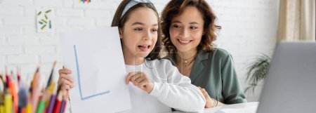 Foto de Chica alegre sosteniendo papel con carta durante la lección de terapia del habla en el ordenador portátil cerca de mamá en casa, pancarta - Imagen libre de derechos