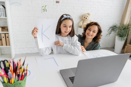 Overjoyed parent looking at laptop near daughter with letter having speech therapy video lesson at home 