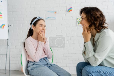 Photo for Smiling preteen pupil touching cheeks during lesson with speech therapist in consulting room - Royalty Free Image