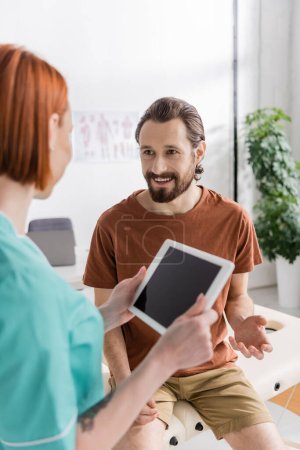 smiling bearded man talking to blurred physiotherapist with digital tablet in rehabilitation center