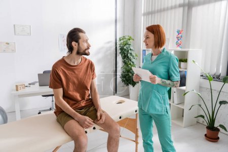 redhead rehabilitation specialist with digital tablet talking to bearded man sitting on massage table in consulting room