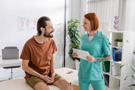 positive bearded man sitting on massage table and talking to redhead physiotherapist with digital tablet in rehab center