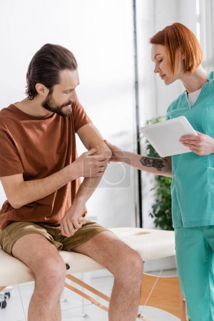 kinésithérapeute rousse avec tablette numérique examinant le coude blessé d'un homme barbu assis sur une table de massage dans une salle de consultation