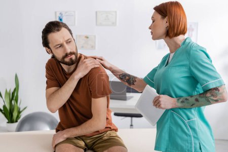Téléchargez les photos : Physiothérapeute tatoué avec tablette numérique touchant l'épaule blessée de l'homme barbu lors d'un rendez-vous dans un centre de réadaptation - en image libre de droit