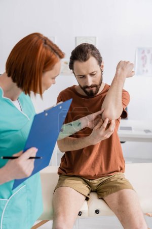 physiotherapist with digital tablet examining painful elbow of bearded man in hospital