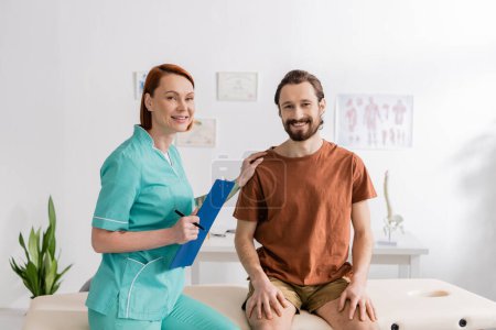 Photo for Cheerful bearded man and redhead physiotherapist with clipboard smiling at camera in consulting room - Royalty Free Image