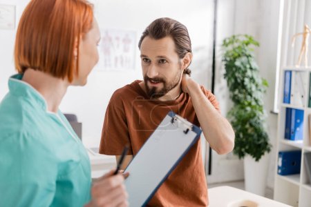 bearded man touching painful neck near blurred physiotherapist with clipboard in consulting room