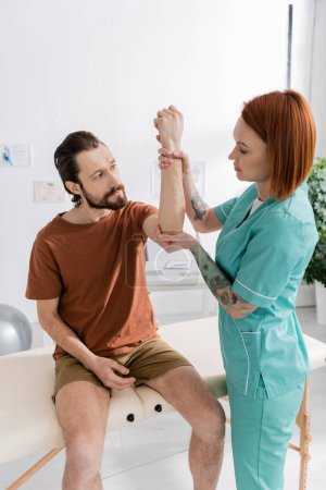 chiropractor examining arm and elbow of injured bearded man sitting on massage table in consulting room