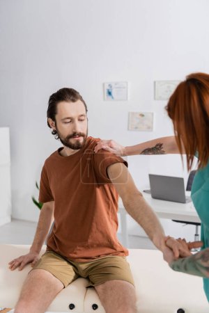 fisioterapeuta pelirroja examinando brazo de hombre barbudo herido en clínica de rehabilitación