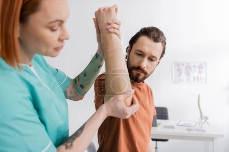 tattooed physiotherapist flexing arm of bearded man while examining injured elbow in consulting room