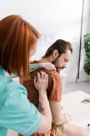 blurred chiropractor examining painful shoulder of injured man in consulting room