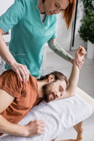 tattooed physiotherapist massaging injured shoulder of bearded man in rehabilitation center