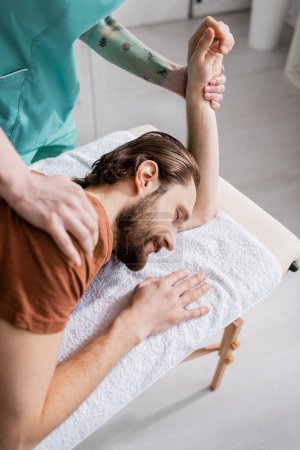 tattooed chiropractor massaging shoulder of injured man during manual therapy in consulting room