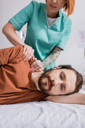 bearded man near physiotherapist massaging injured shoulder during treatment in rehabilitation center