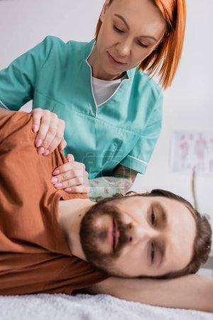 pelirroja terapeuta manual masajeando doloroso hombro de barbudo hombre en centro de rehabilitación