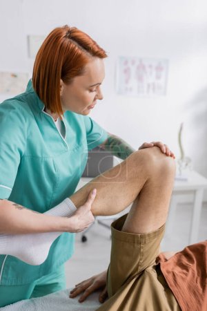 Rotschopf-Physiotherapeut beugt verletztem Mann bei Reha-Therapie im Sprechzimmer Bein