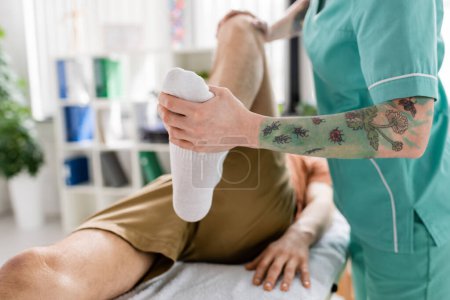 Téléchargez les photos : Vue recadrée du chiropraticien tatoué flexion jambe du patient pendant le traitement dans le centre de réadaptation - en image libre de droit