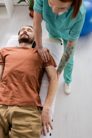 Hochwinkelaufnahme eines bärtigen Mannes, der in der Nähe eines Physiotherapeuten liegt und im Erholungszentrum Arm- und Schultermassage macht