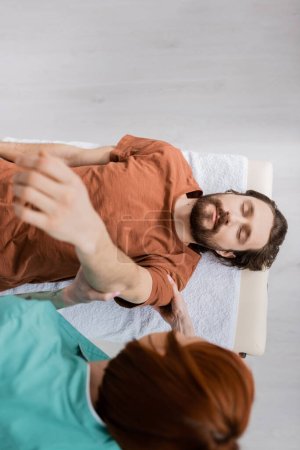 top view of blurred chiropractor stretching injured arm of bearded man with closed on massage table in consulting room