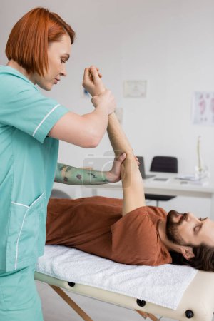 kinésithérapeute rousse étirant le bras douloureux de l'homme barbu couché sur la table de massage dans le centre de réadaptation