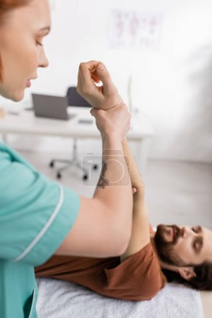 physiotherapist stretching injured arm of blurred man during rehabilitation in hospital