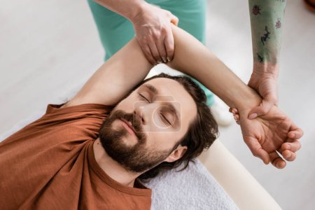 Téléchargez les photos : Vue de dessus de l'homme barbu avec les yeux fermés près du physiothérapeute faisant massage des bras pendant la réadaptation à la clinique - en image libre de droit