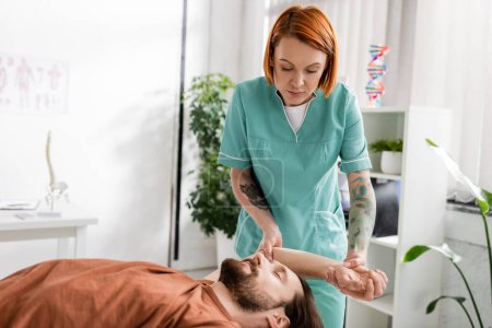 tattooed manual therapist massaging painful arm of bearded man in rehabilitation center