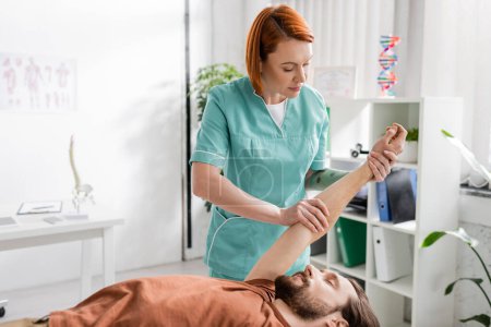 redhead chiropractor stretching arm of injured man while doing pain relief massage in consulting room