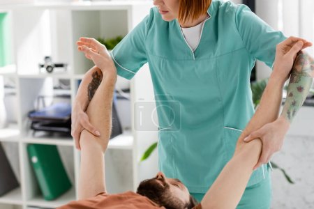 Manuelle Therapeutin streckt Arme eines bärtigen Mannes während der Rehabilitation in der Klinik