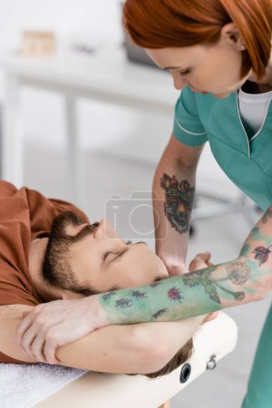 tattooed osteopath massaging injured arm of bearded man in recovery center