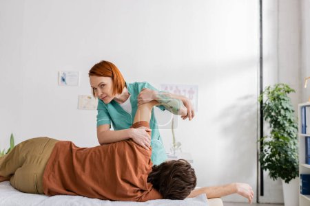redhead chiropractor looking at camera while flexing injured arm of man during pain relief massage in consulting room