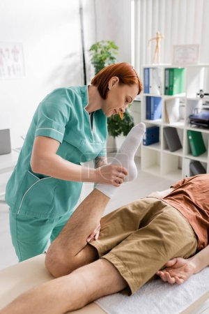 rudowłosa osteopatka napinająca nogę mężczyzny leżącego na stole do masażu w centrum rehabilitacyjnym