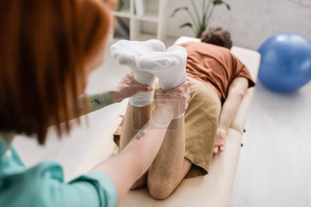 Téléchargez les photos : Physiothérapeute flou jambes fléchissantes de l'homme blessé lors de la manipulation de récupération sur table de massage à la clinique - en image libre de droit