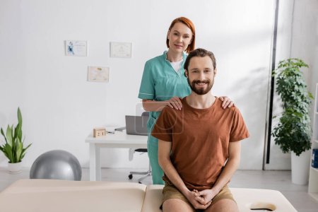 Foto de Fisioterapeuta feliz tocando hombros de hombre barbudo sentado en la mesa de masaje en la sala de consulta - Imagen libre de derechos