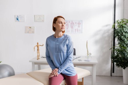 Photo for Redhead woman sitting on massage table in rehab center and looking away - Royalty Free Image