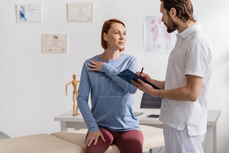 redhead woman touching painful shoulder near physiotherapist writing prescription on clipboard in consulting room