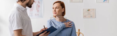 woman touching injured shoulder near physiotherapist writing diagnosis on clipboard in consulting room, banner