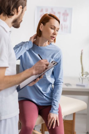 woman touching injured neck near chiropractor writing diagnosis on clipboard in consulting room