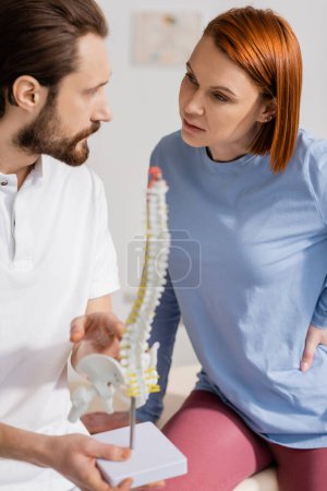 bearded osteopath holding spine woman and talking to redhead woman in consulting room