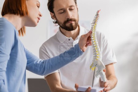 redhead woman touching spine model near bearded physiotherapist in consulting room