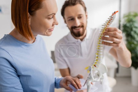 smiling chiropractor showing spine model to redhead woman during appointment in consulting room