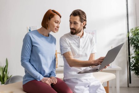 Téléchargez les photos : Physiothérapeute barbu avec ordinateur portable parlant à une femme souriante lors d'un rendez-vous dans un centre de réadaptation - en image libre de droit