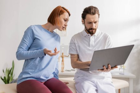 physiothérapeute barbu à l'aide d'un ordinateur portable près rousse femme pointant avec la main dans la salle de consultation